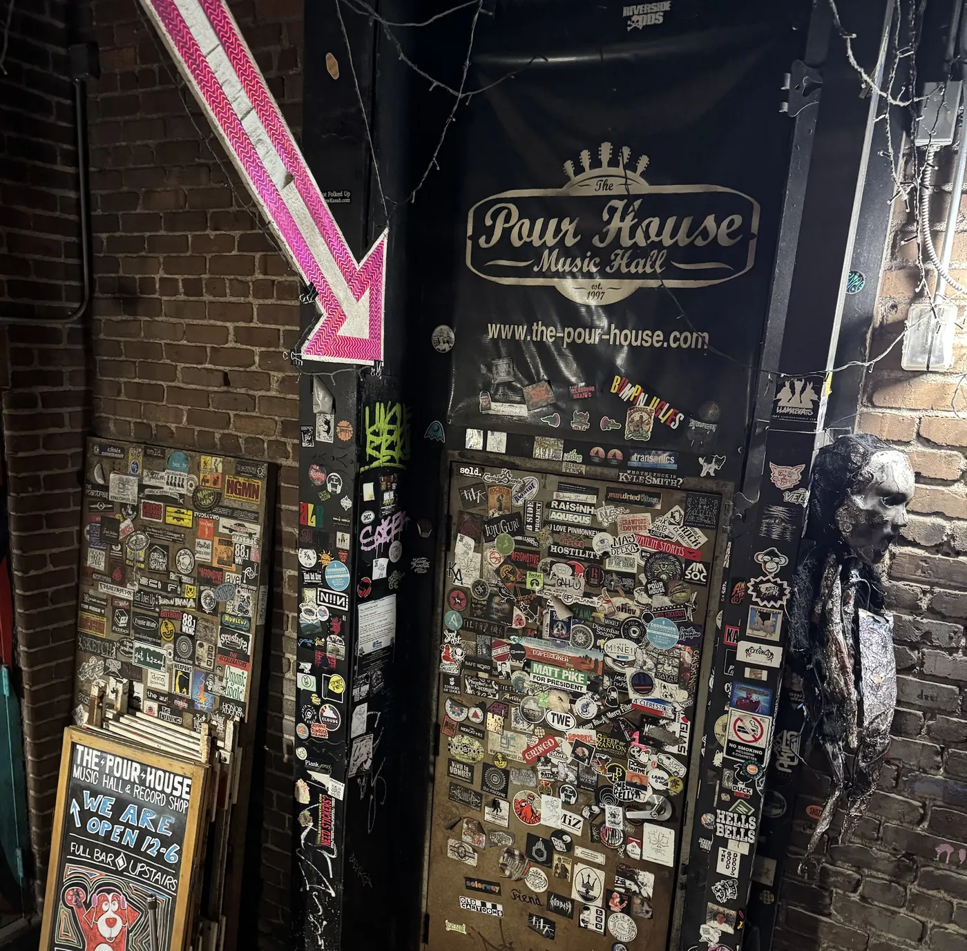 The Pour House Music Hall door in a brick alley, covered in stickers, under a pink arrow sign