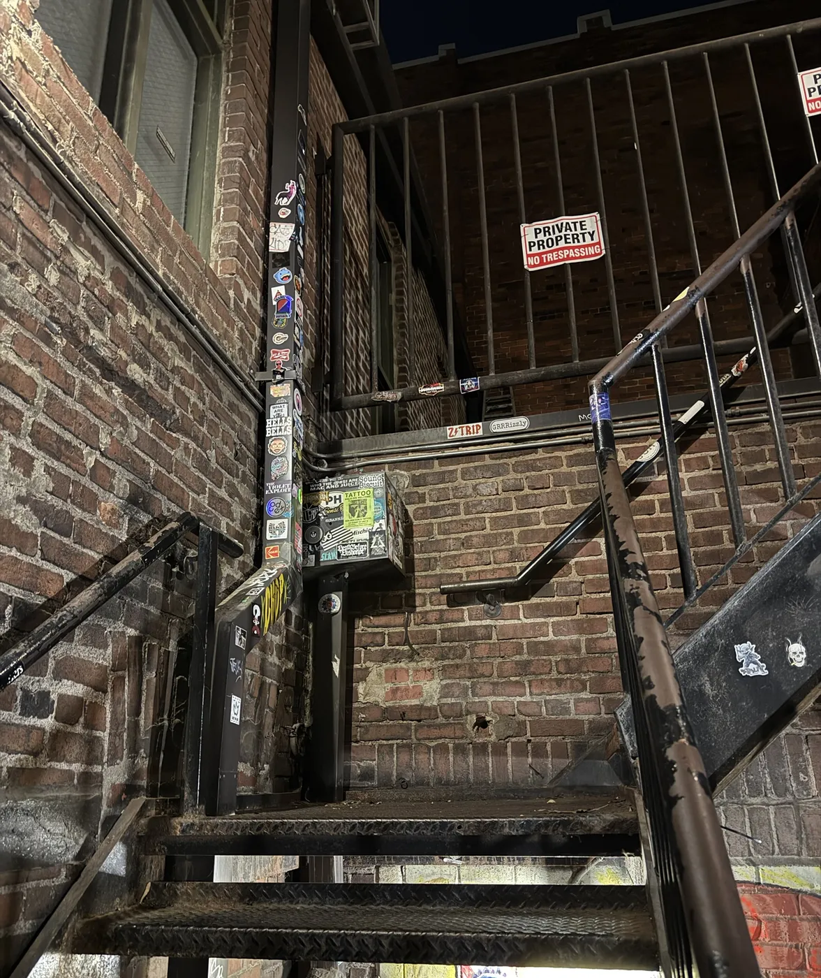 Steel stairs up a brick wall, the rails and drainpipe covered in stickers