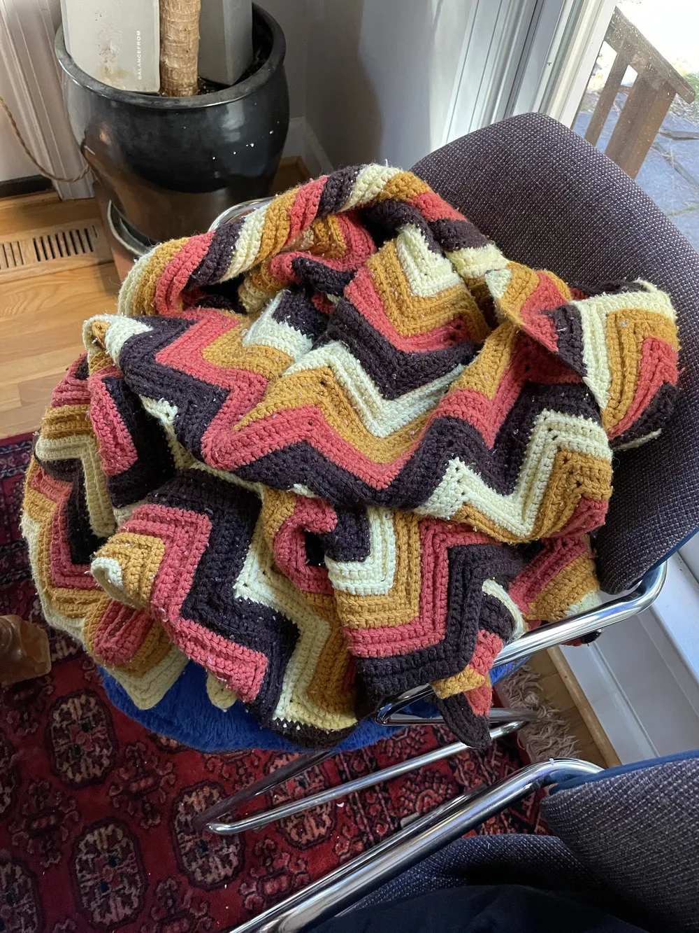 A chevron crocheted blanket in brown, coral, mustard and cream piled in a lump on a chrome chair, with a dog sleeping underneath